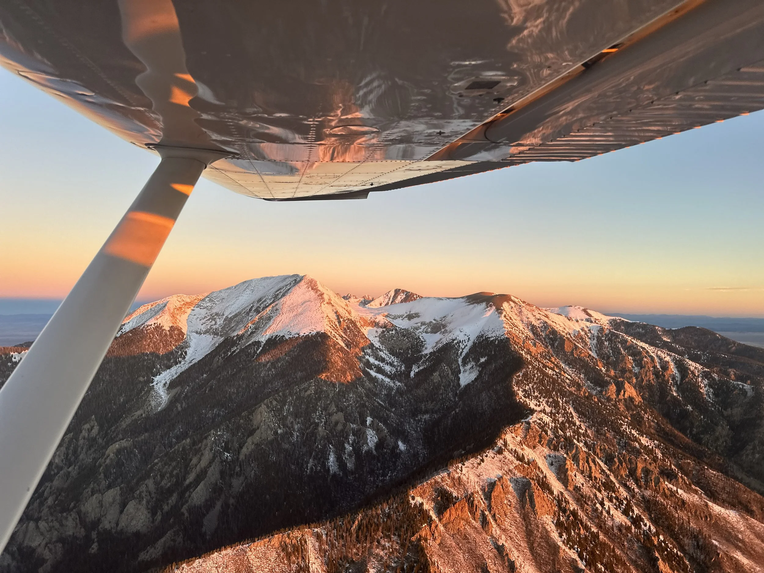 Aircraft wing view over snow-capped mountains at sunset