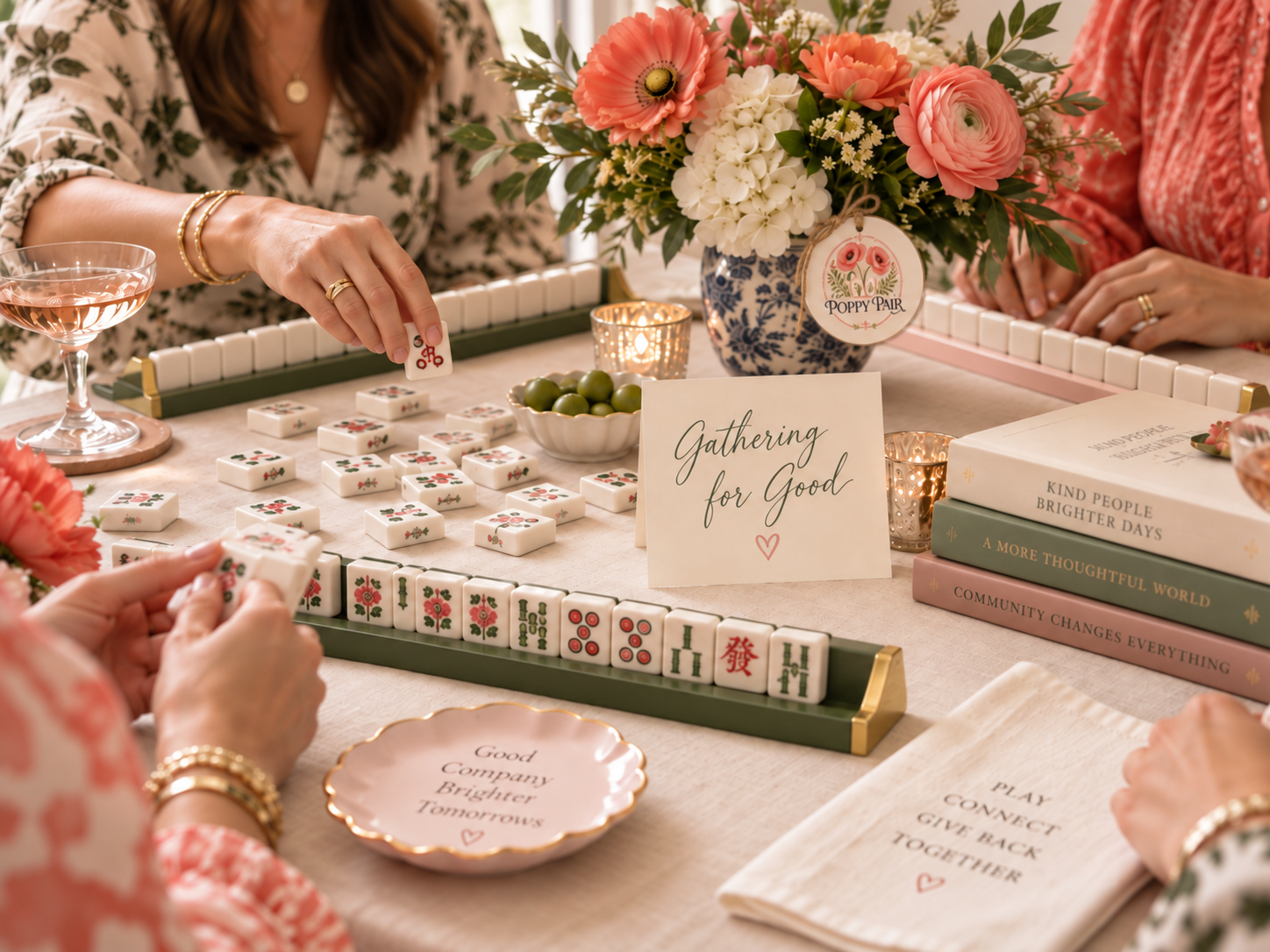 Women playing American Mah Jongg at a styled table with coral flowers, The Poppy Pair logo, and a “Gathering for Good” card.
