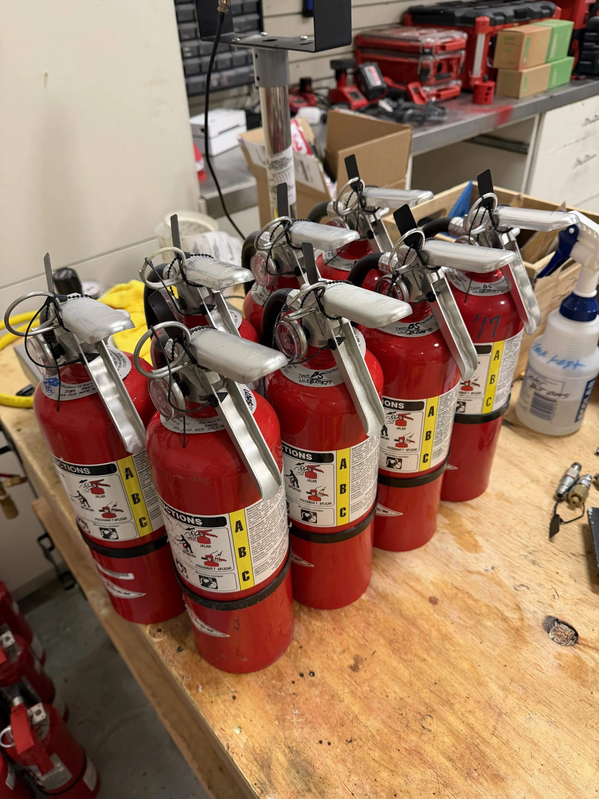 A row of red fire extinguishers with black, yellow, and white labels, placed on a wooden workbench in a workshop. In the background, there are various tools and boxes, including a spray bottle on the right.