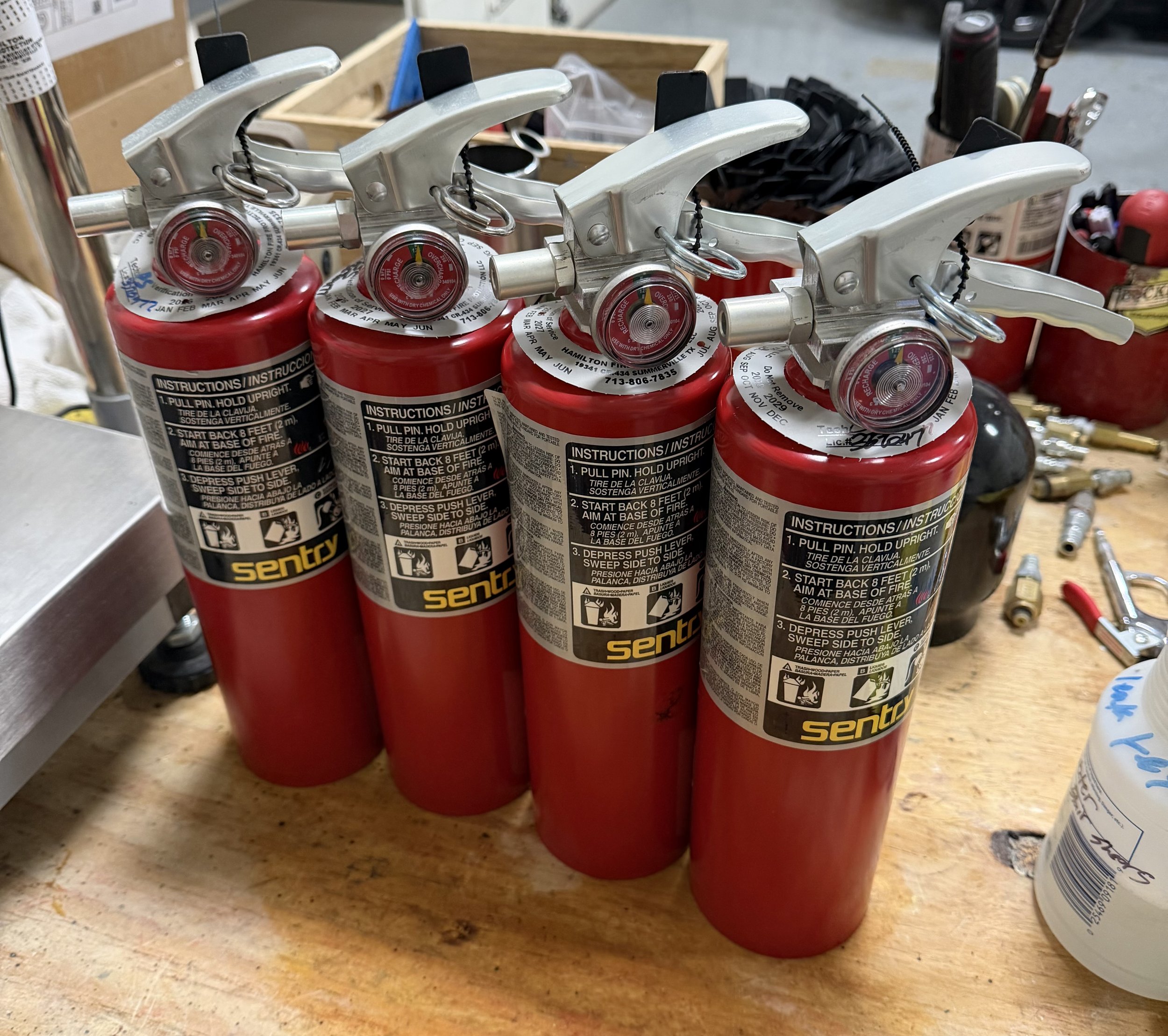 A row of four red fire extinguishers with black and silver nozzles and instructions printed on the labels, placed on a wooden surface in a workshop or store environment.