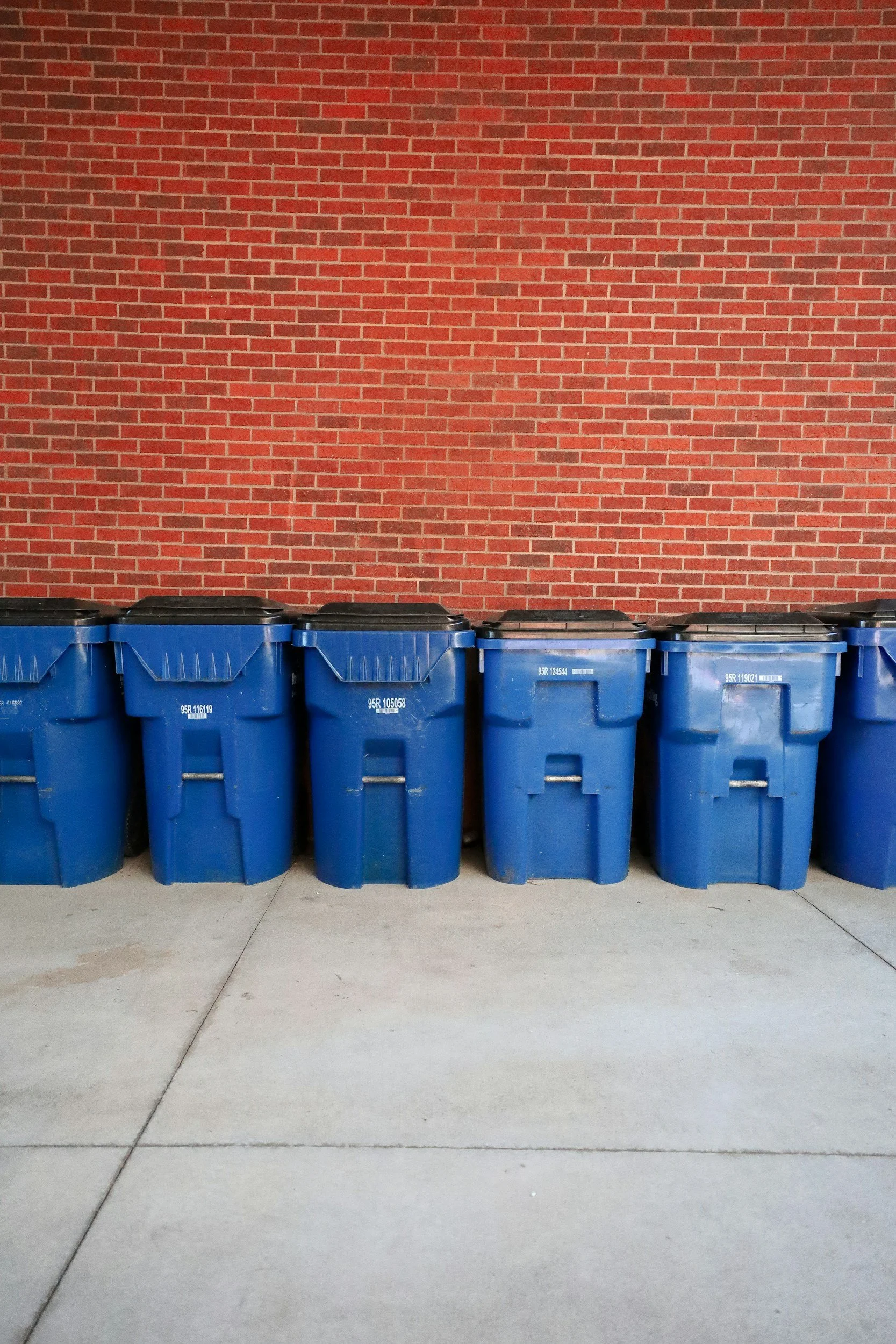 A row of blue trash and recycling bins lined up against a red brick wall on a concrete sidewalk.