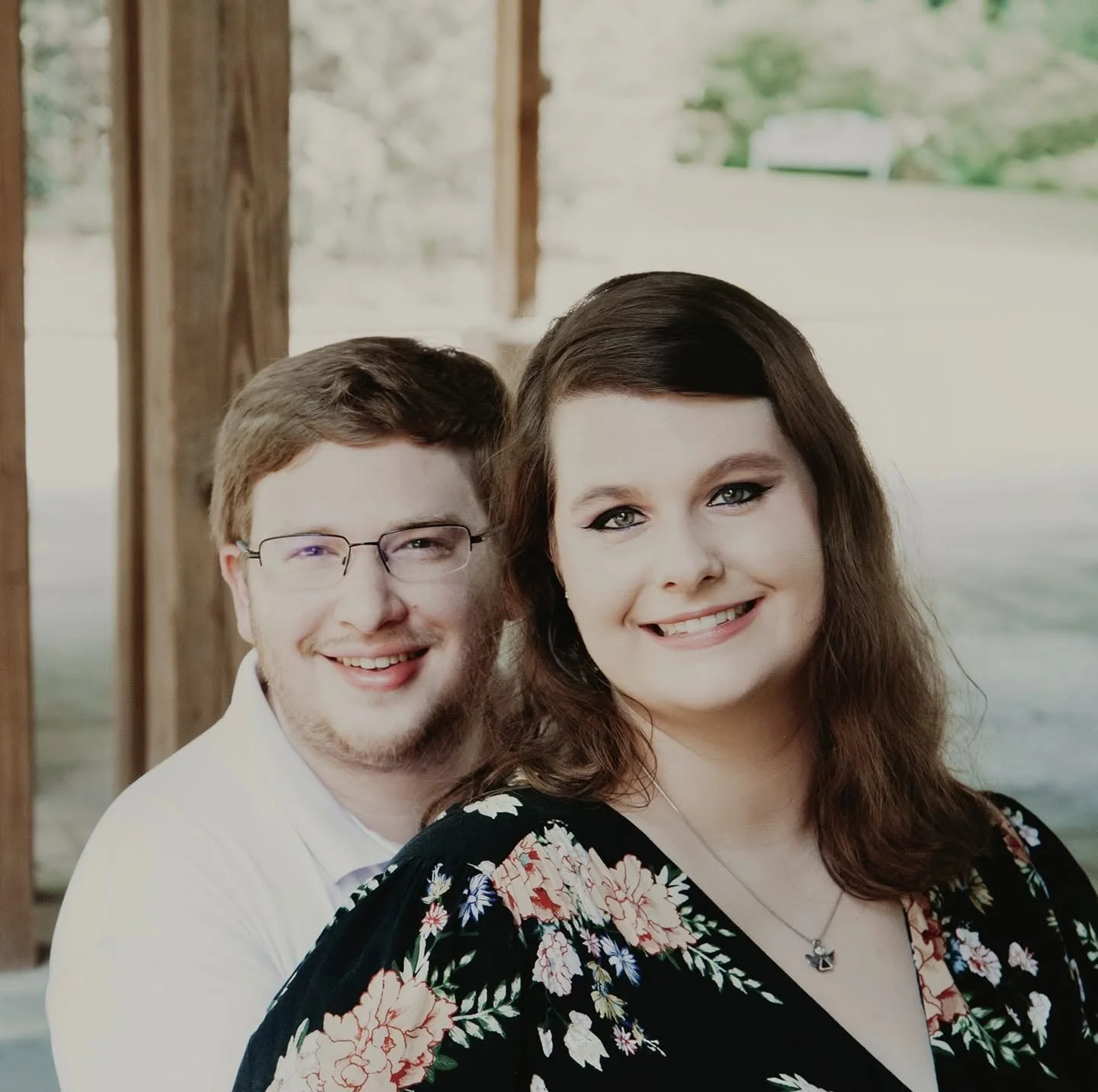 A smiling man with glasses and a woman with long brown hair, both dressed nicely, standing outdoors near wooden posts.