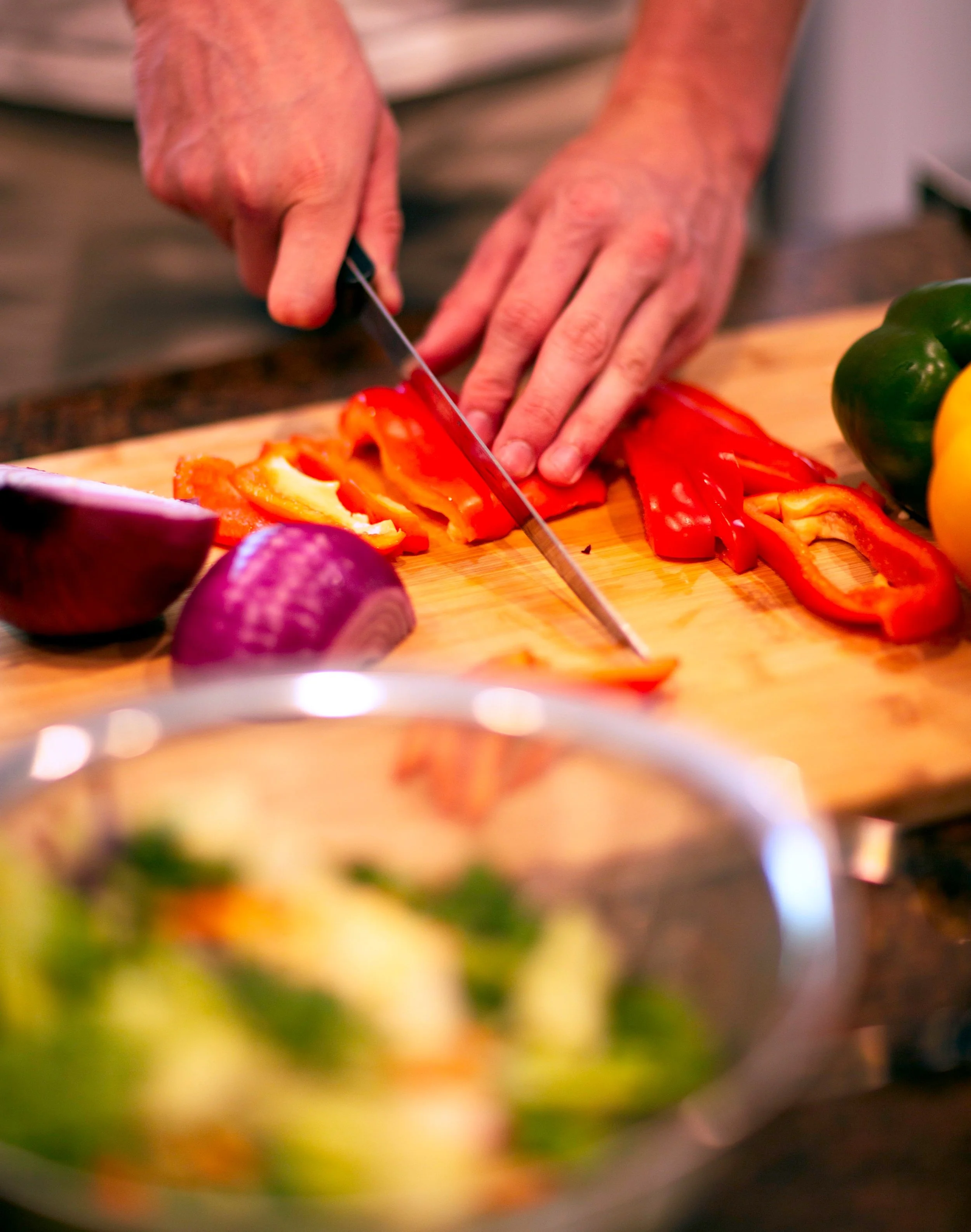 Person slicing red and yellow bell peppers on a wooden cutting board, with other vegetables like red onion, green, and yellow bell peppers nearby.