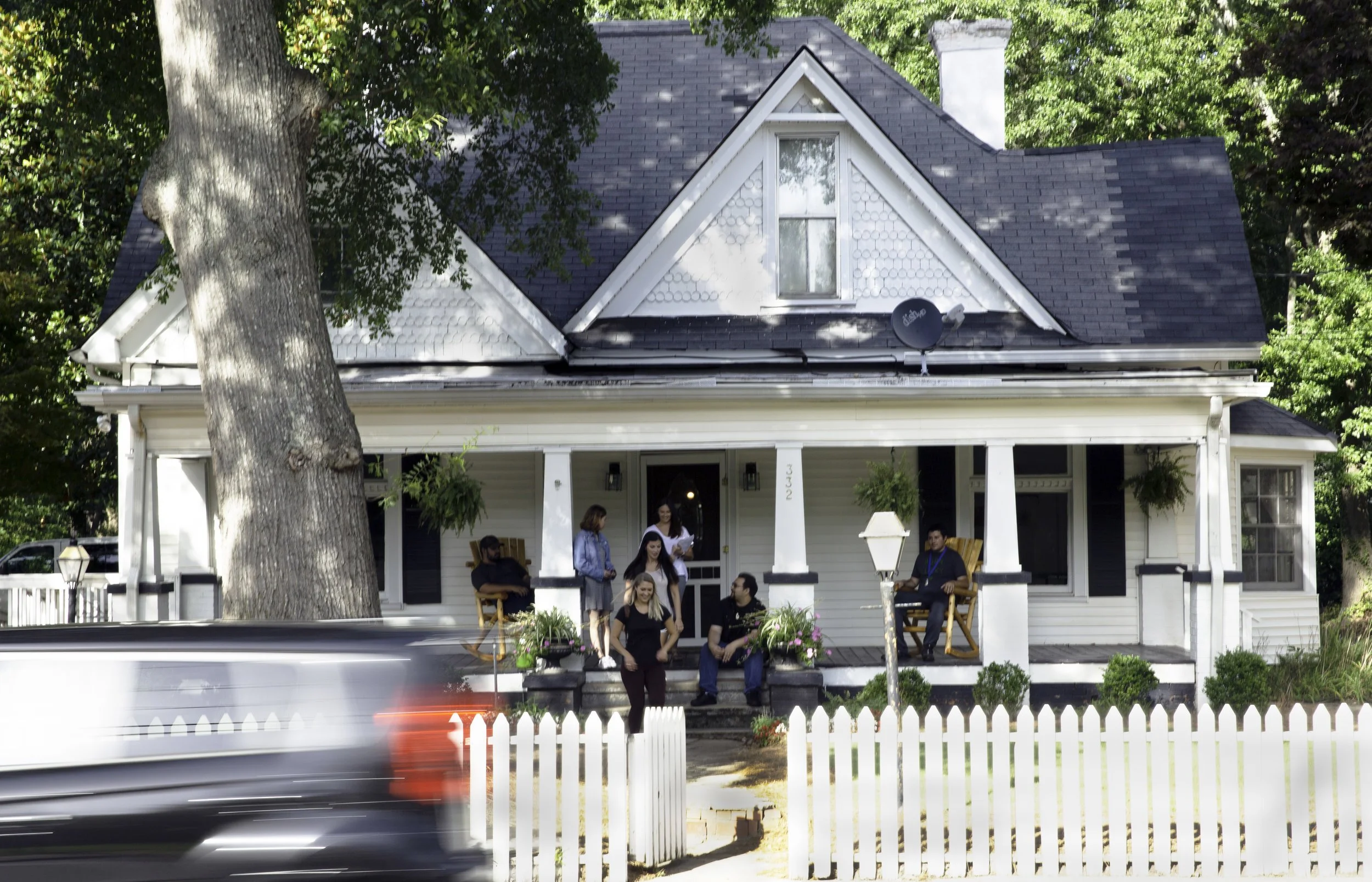 A group of people on the front porch of a white house with black shutters, surrounded by trees and a white picket fence. A car passes by in the foreground.