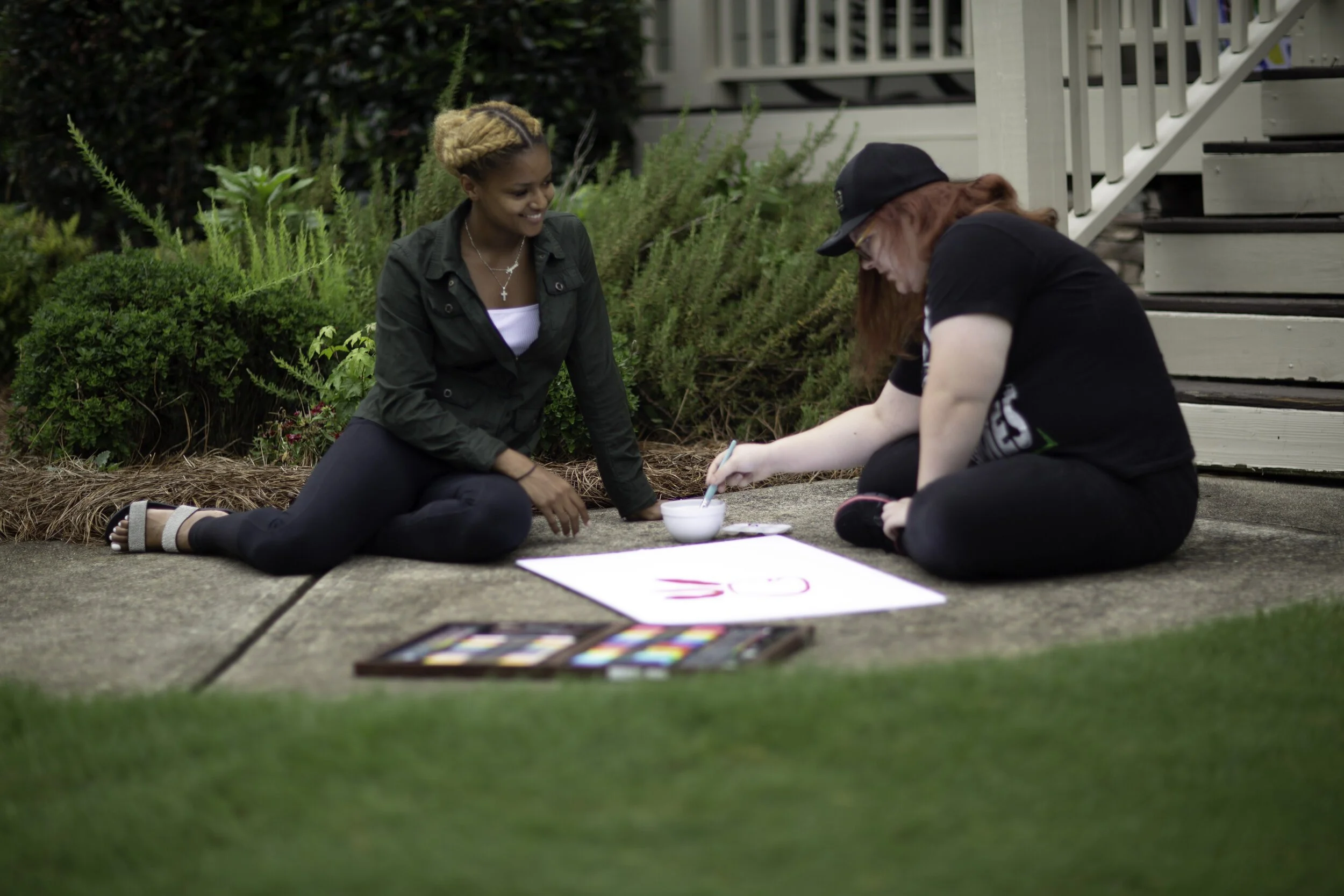 Two women are sitting on a concrete sidewalk outside, drawing on a large white poster with pink paint. One woman has curly red hair, is wearing a black cap and black clothes, and is using a paintbrush. The other woman has blonde hair tied back, is smiling, and is wearing a dark green jacket and black pants. A colorful palette and a bowl of water are on the ground near them.