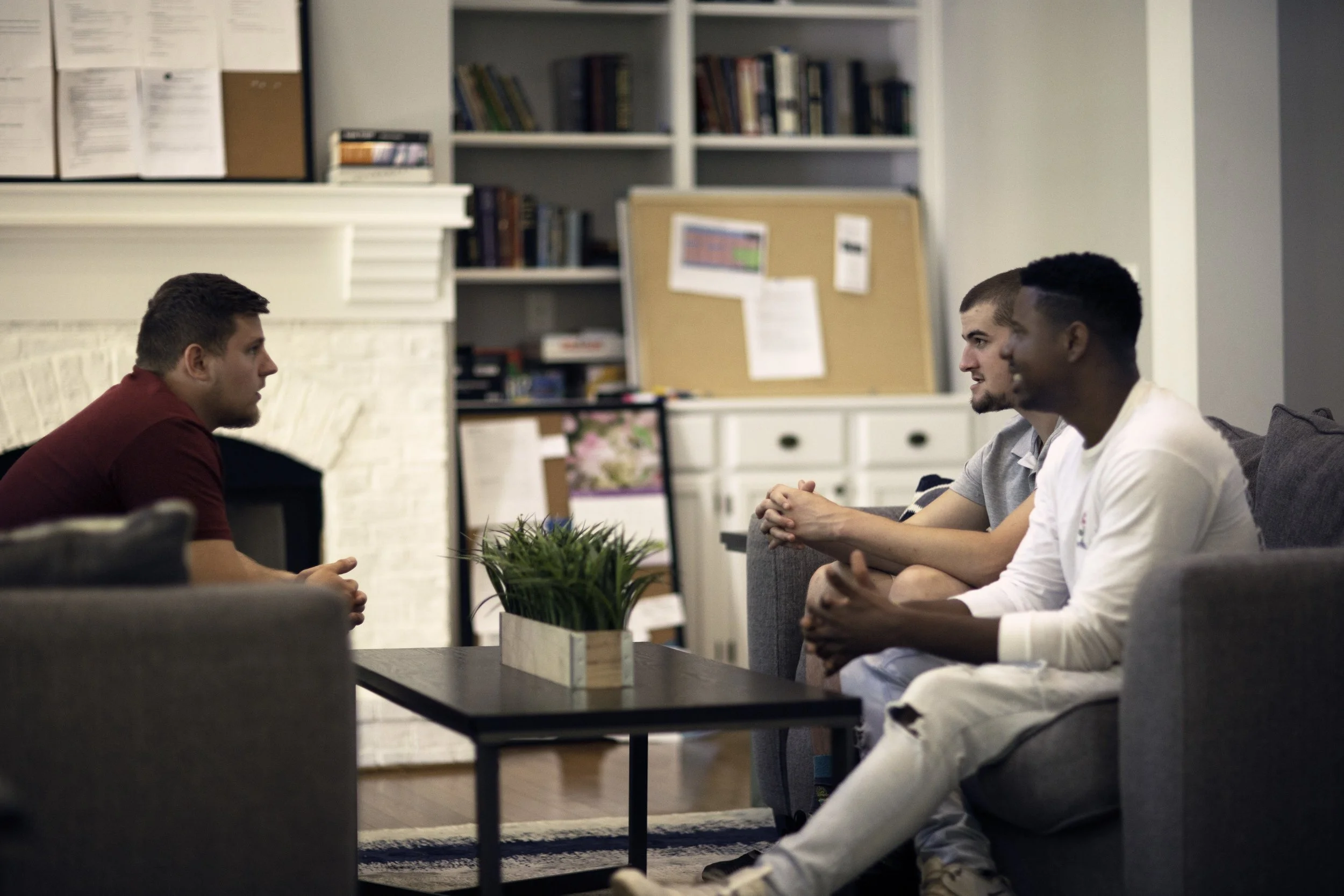 Three men sit on a couch across from a man sitting on an armchair, engaged in conversation inside a cozy living room.
