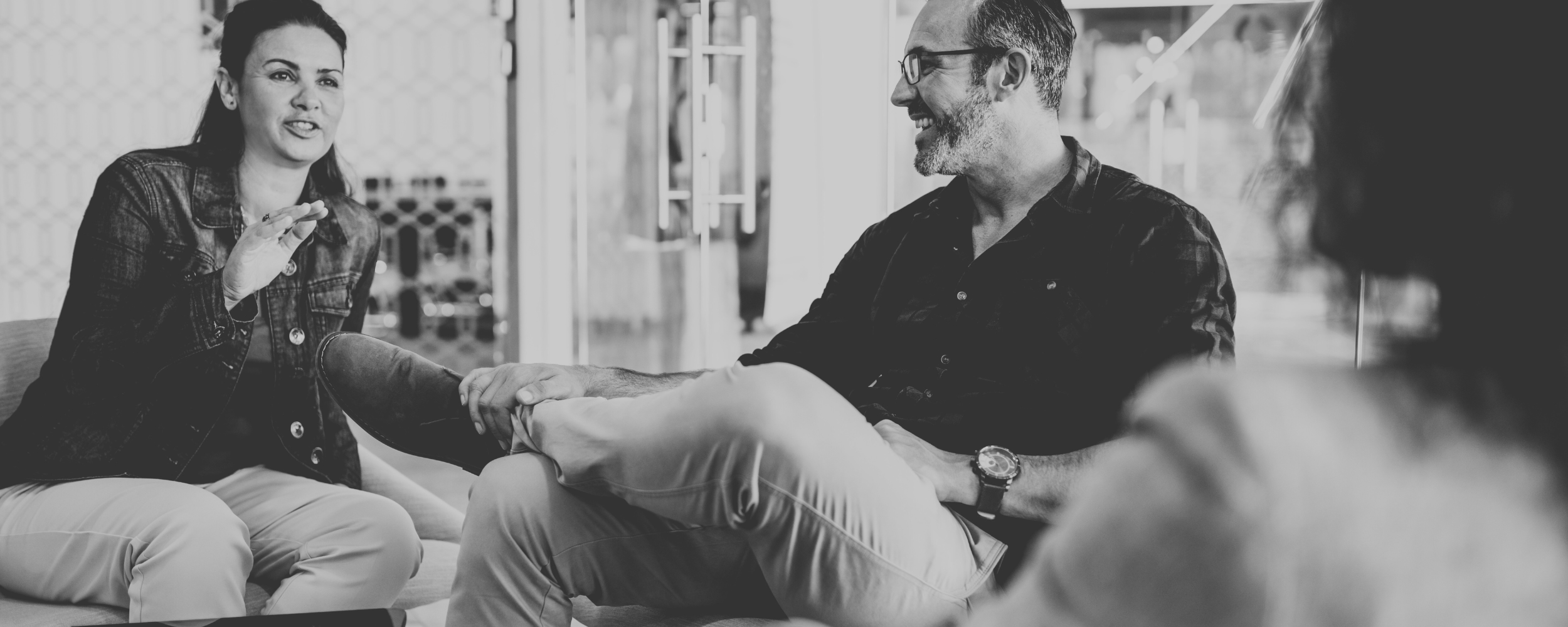 A group of three people sitting and having a conversation in an indoor setting. The woman on the left is speaking, and a man on the right is smiling and holding her foot. The third person is partially visible on the right side.