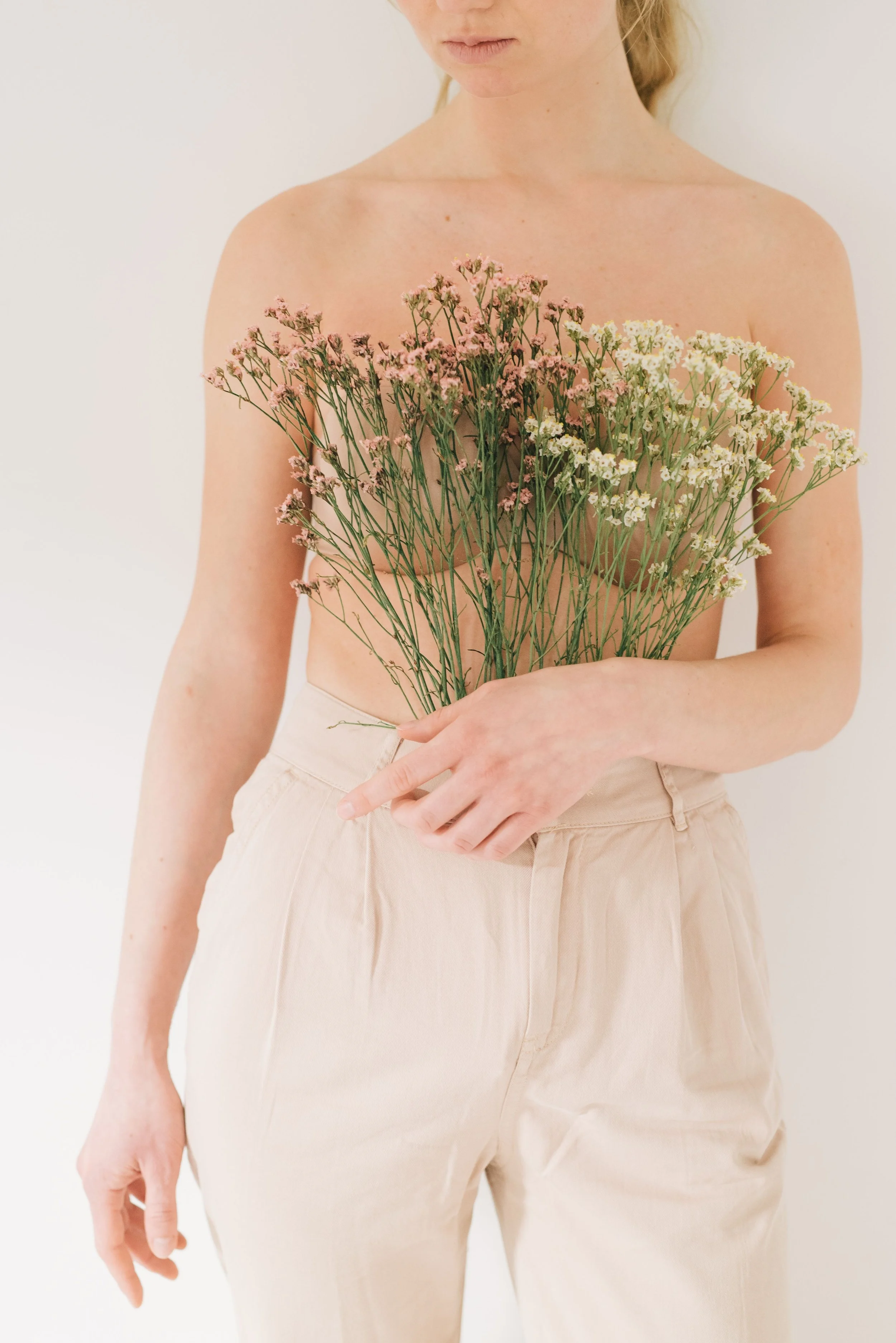 A woman with light skin and blonde hair holding a bouquet of pink and white flowers, wearing beige pants against a plain white background.