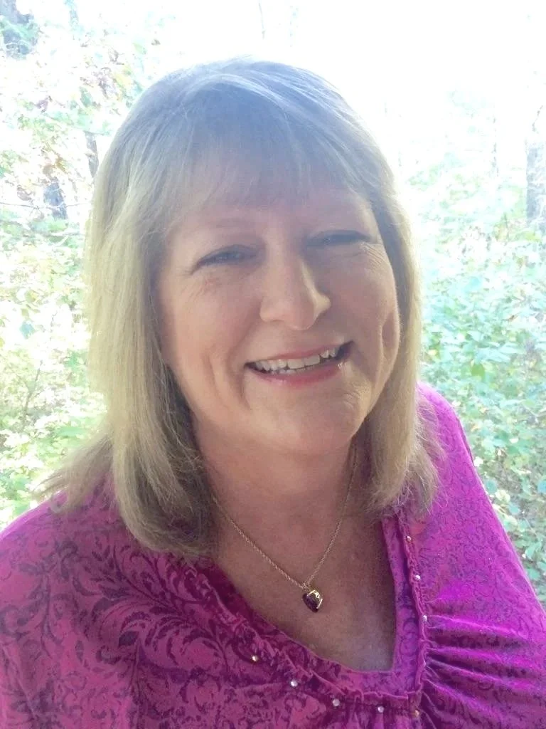 A middle-aged woman with shoulder-length blonde hair smiling, wearing a purple top with a pattern and a heart-shaped necklace, standing outdoors with green foliage in the background.
