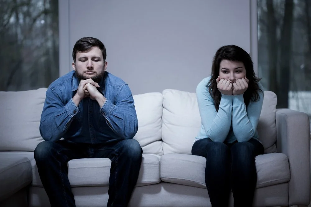 A man and a woman sit on a white couch inside a room with large windows showing trees outside. The man has brown hair and is wearing a denim shirt, sitting with his hands clasped under his chin and eyes closed. The woman has dark hair, wearing a light blue sweater, sitting with her elbows on her knees, resting her face in her hands, and looking down thoughtfully.