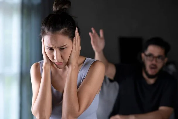 A woman covering her ears in distress as a man in the background yells at her.