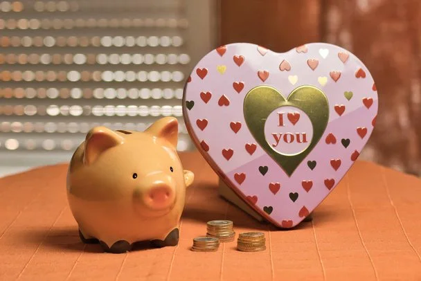 A piggy bank and a heart-shaped box with a gold and green heart and the words 'I love you' on it, alongside some coins on a wooden surface.