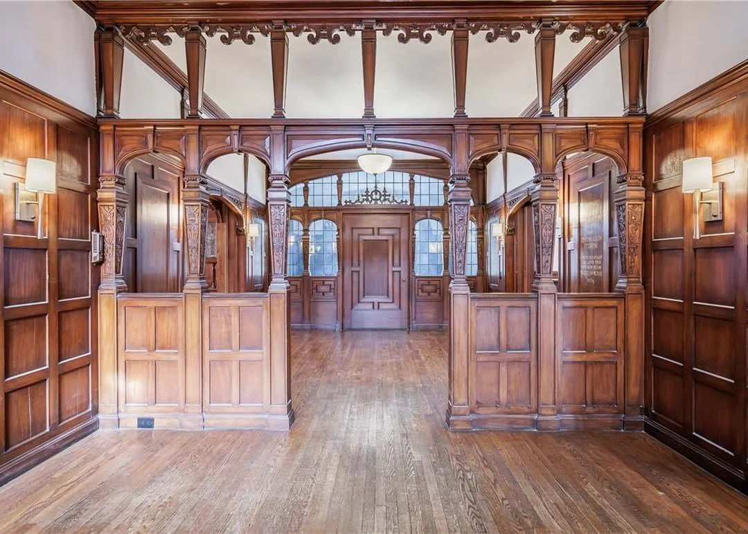 An interior view of a wood-paneled room with a decorative wooden archway barrier, wood flooring, and wall sconces lighting the space. There are windows with a rounded top and a door in the background.