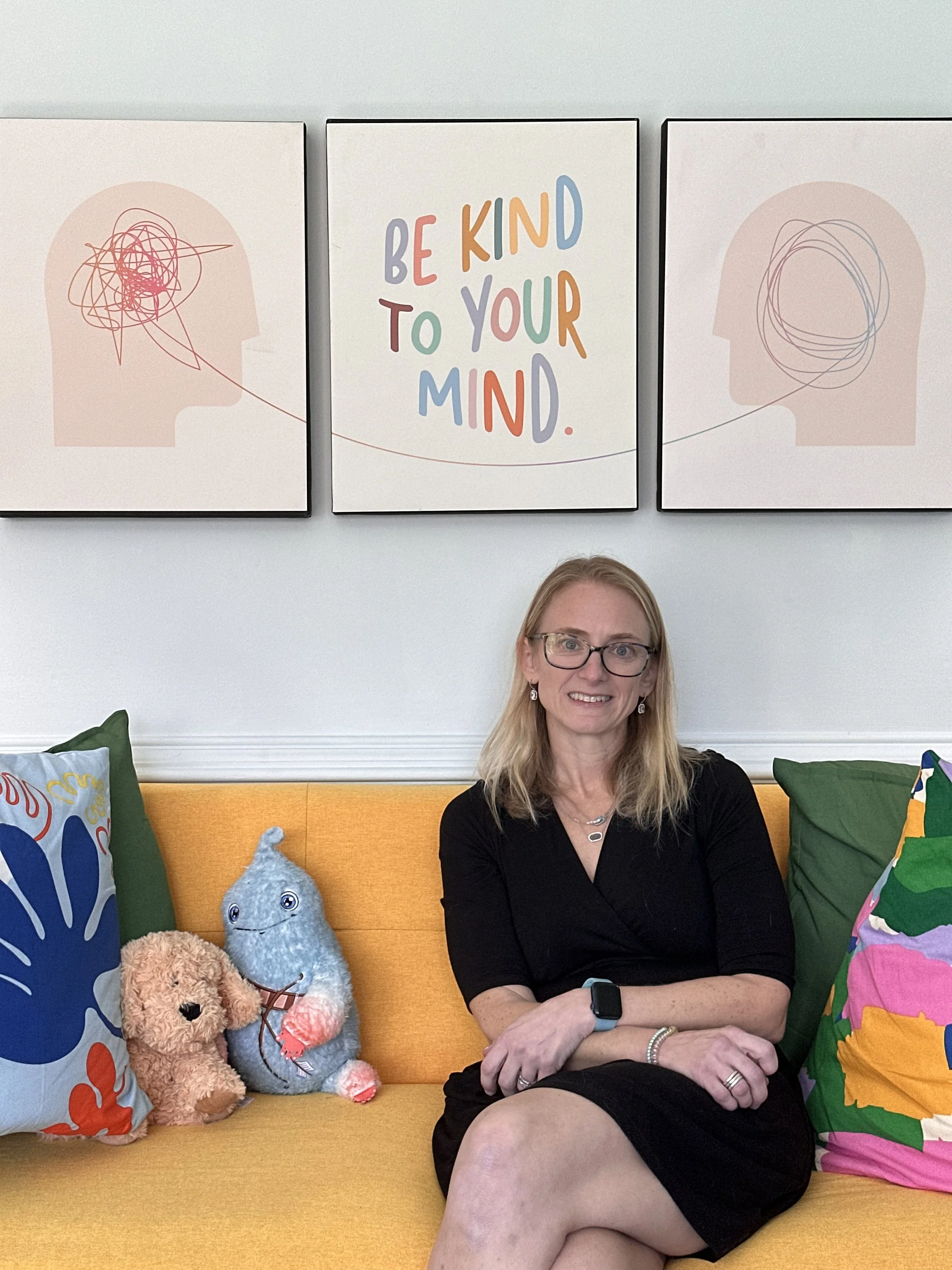 A woman with glasses sitting on a yellow couch, smiling at the camera. Behind her are colorful pillows, plush toys, and artwork on the wall including a sign that says, "Be kind to your mind."