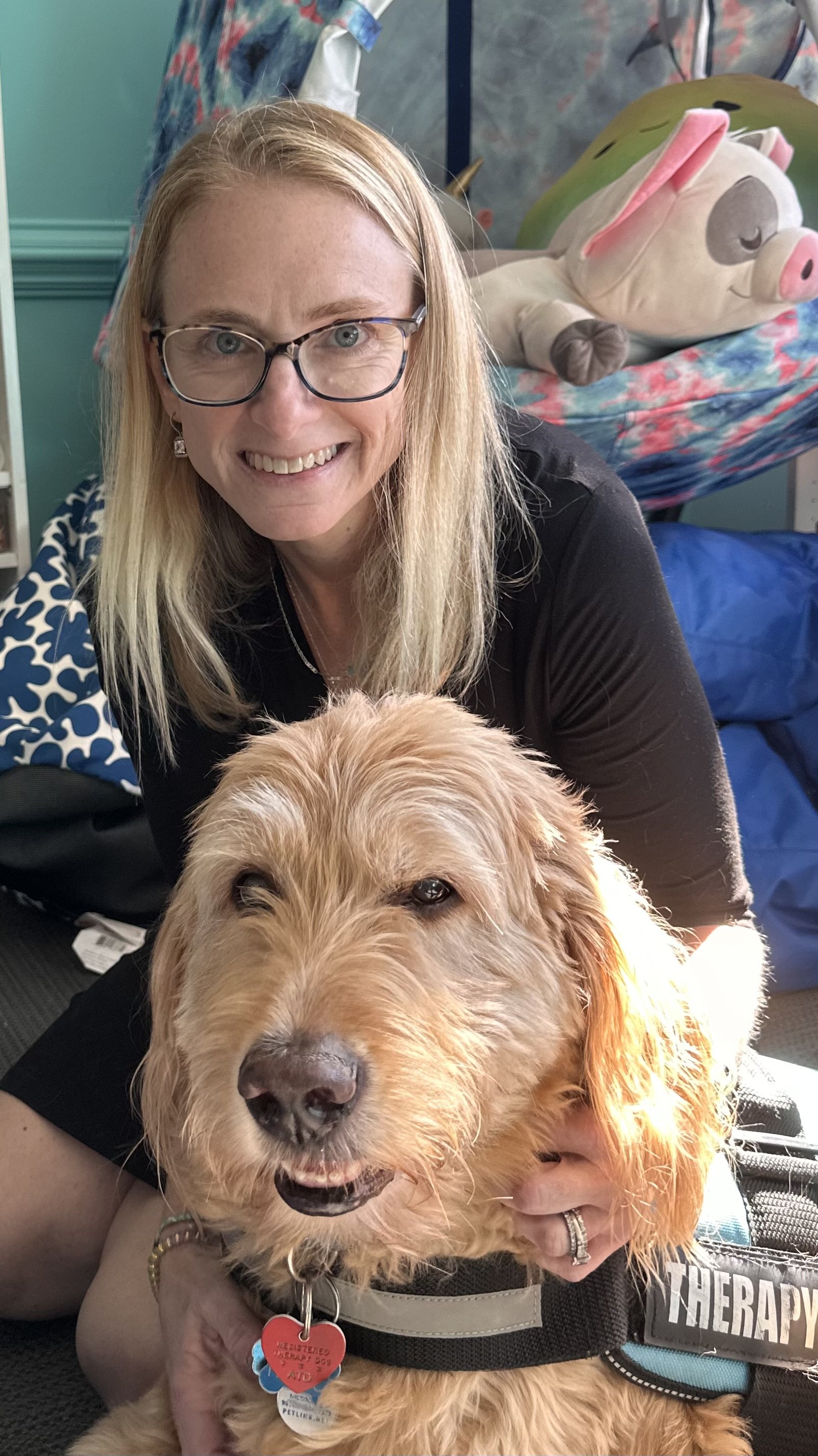 A woman with blonde hair, glasses, and a black shirt smiling, leaning over a golden retriever dog with a leash and tags, inside a room with a plush pig pillow in the background.