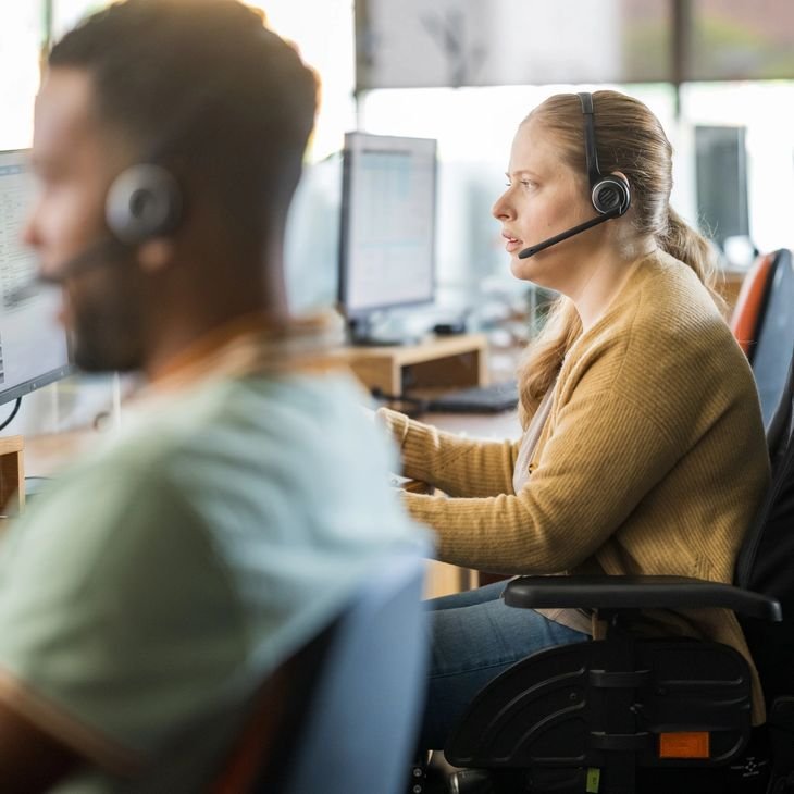 Woman in an office wearing a headset, working at a computer, with another person in the foreground also wearing a headset.