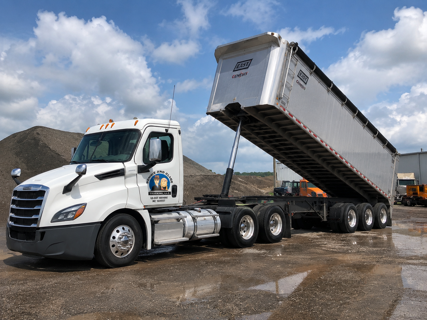 A white semi-truck with a raised trailer in a dirt lot, with a logo on the door reading "Donnie and Claudia Trucking LLC," and a clear blue sky with some clouds.