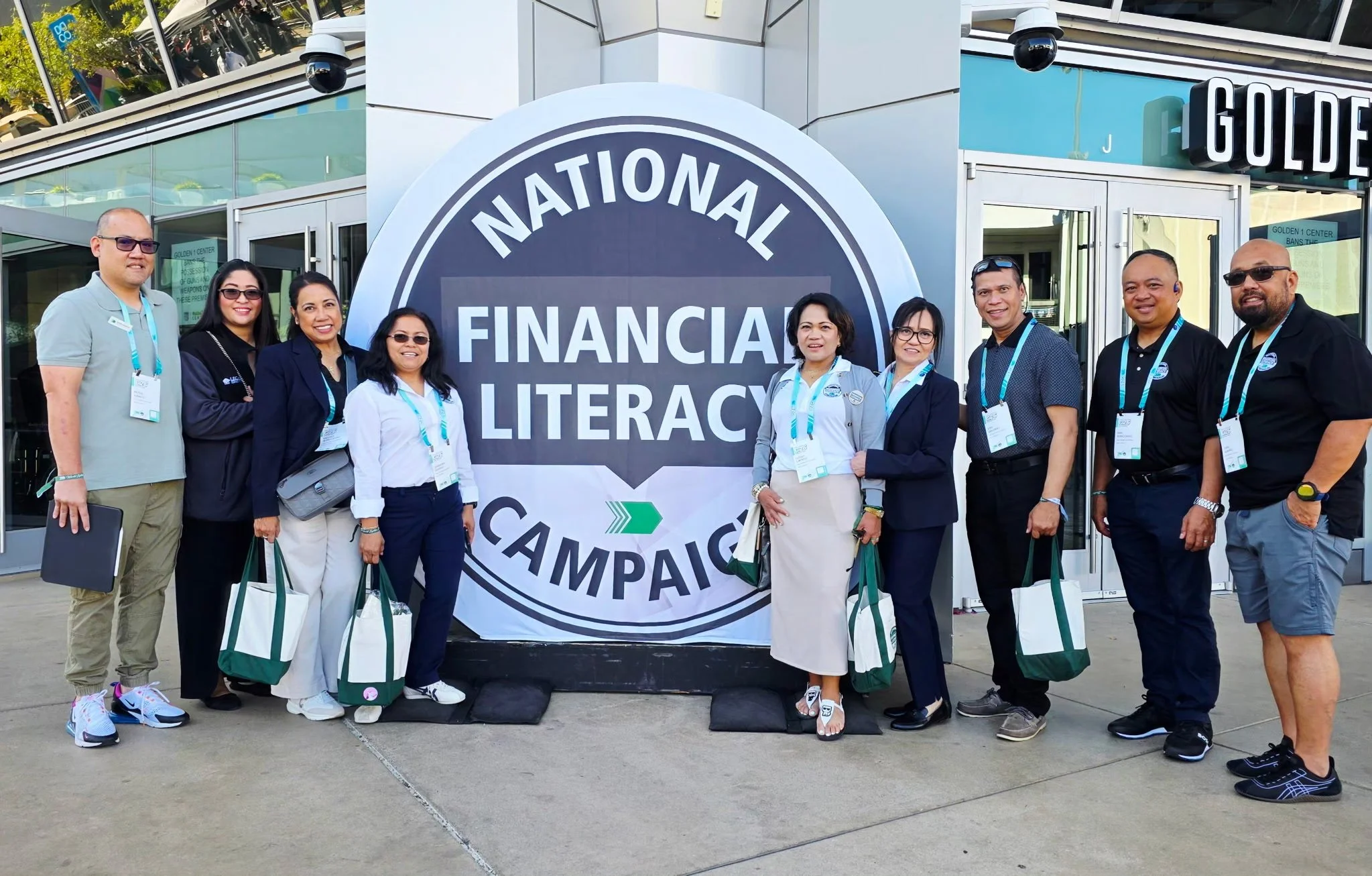 Group of diverse people standing in front of a large sign that reads 'National Financial Literacy Campaign.' They are outdoors, smiling, many wearing conference badges, and holding tote bags. The background shows a glass building entrance.