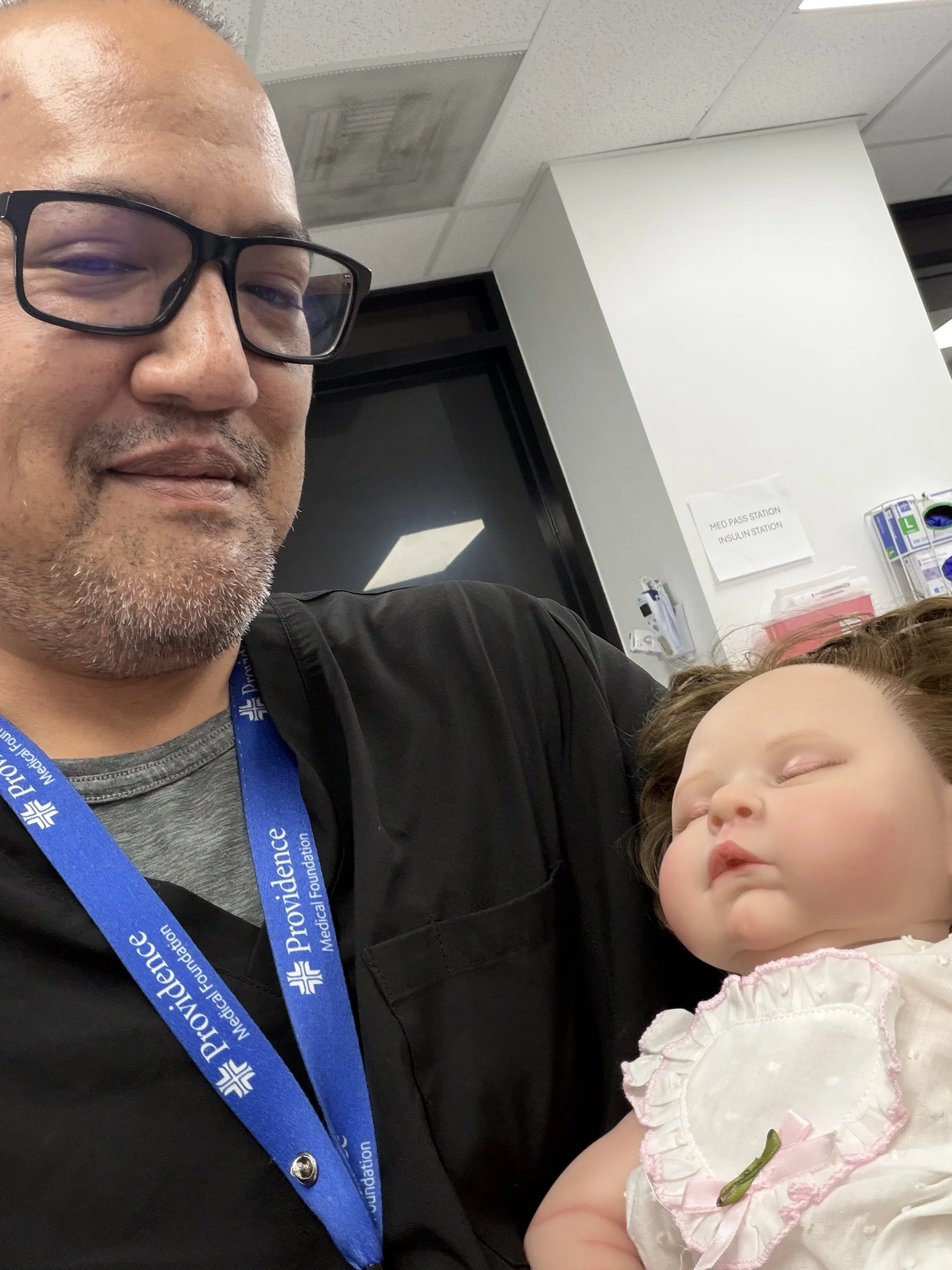A man with glasses and a grey beard wearing a black jacket and a blue badge, holding a sleeping baby girl dressed in a white dress with pink bows, pink frills, and a green flower, inside a medical facility.