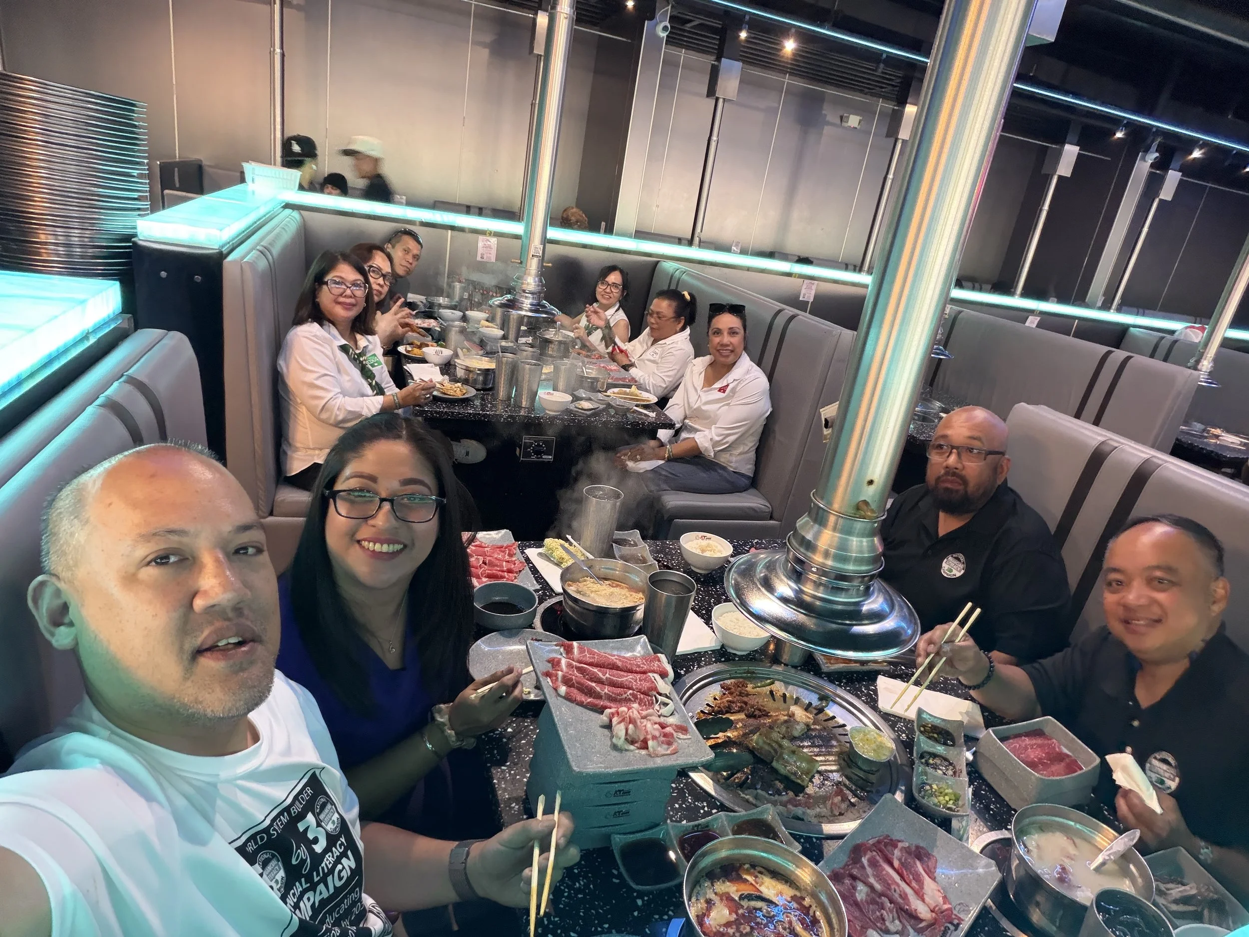 Group of people dining at a restaurant, enjoying a hot pot meal with various ingredients and dishes on the table, including meats, vegetables, and dipping sauces, with a metal ventilation pipe overhead.