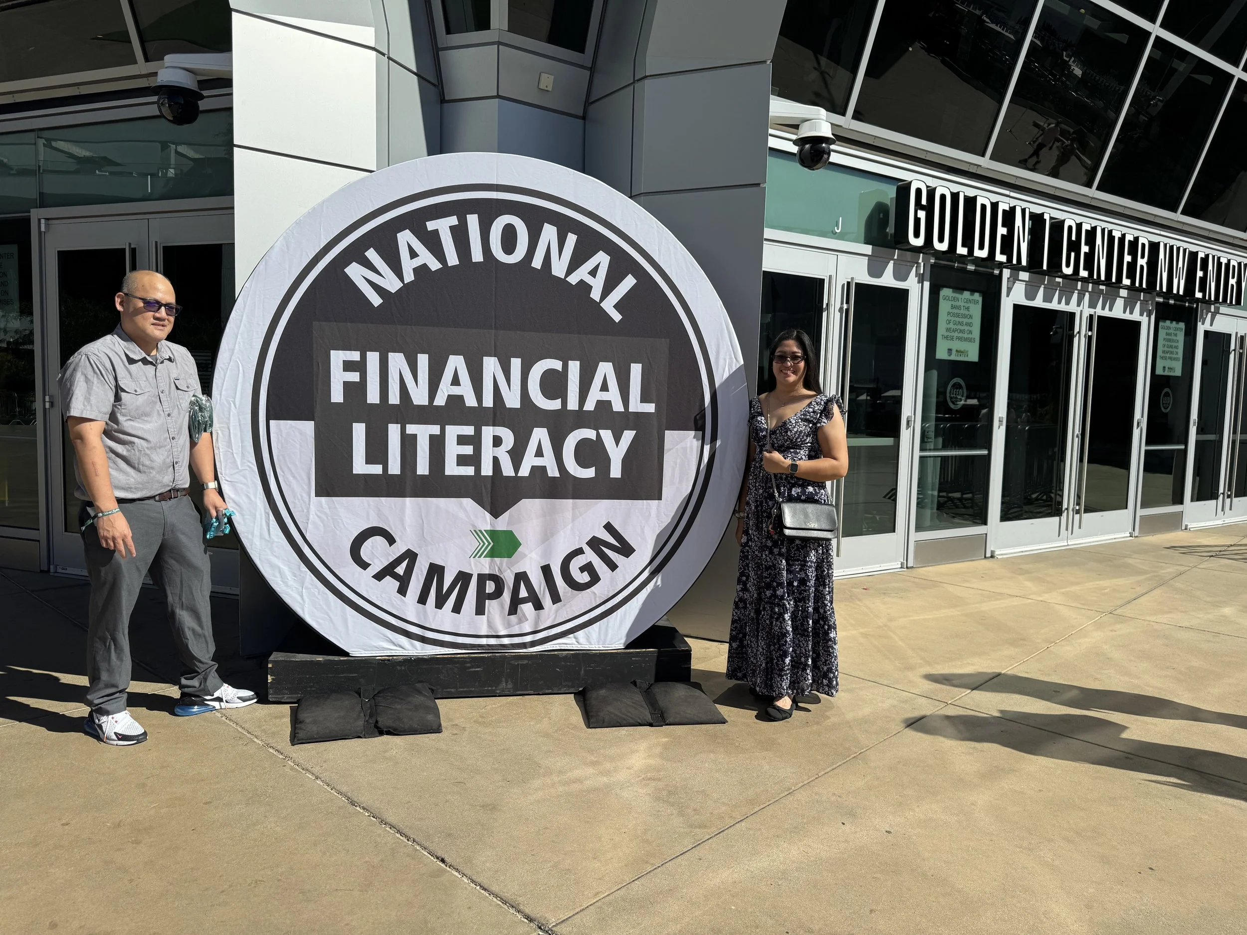 Two people standing next to a large foldable banner that reads 'National Financial Literacy Campaign.' They are outdoors in front of a modern building with glass windows and an entrance labeled 'Golden Center NW Entrance.' One man is wearing sunglasses, a gray shirt, and gray pants; the woman is wearing a black and white dress and has a black purse.