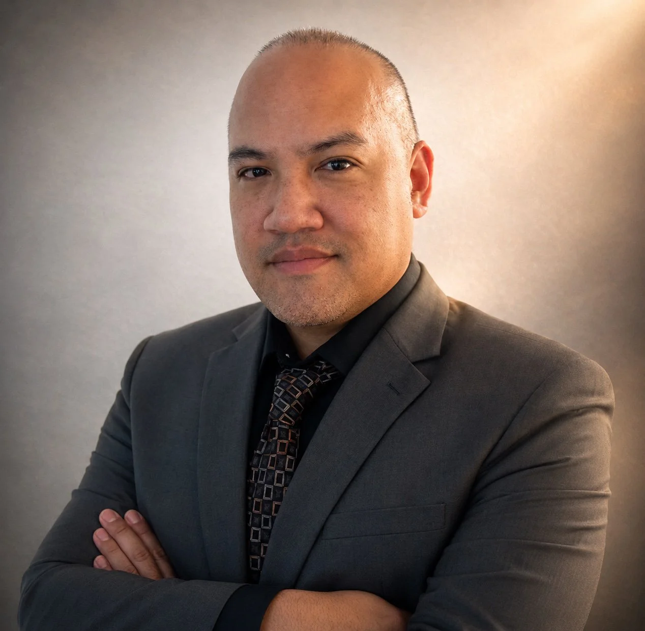 A man in a dark grey suit, black shirt, and patterned tie with crossed arms, standing against a plain neutral background.