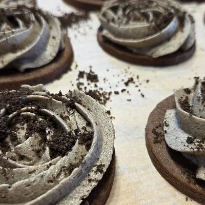 Chocolate cookies topped with cookies‑and‑cream frosting piped in rose‑shaped swirls, with dark crumbs sprinkled across the frosting and surface.