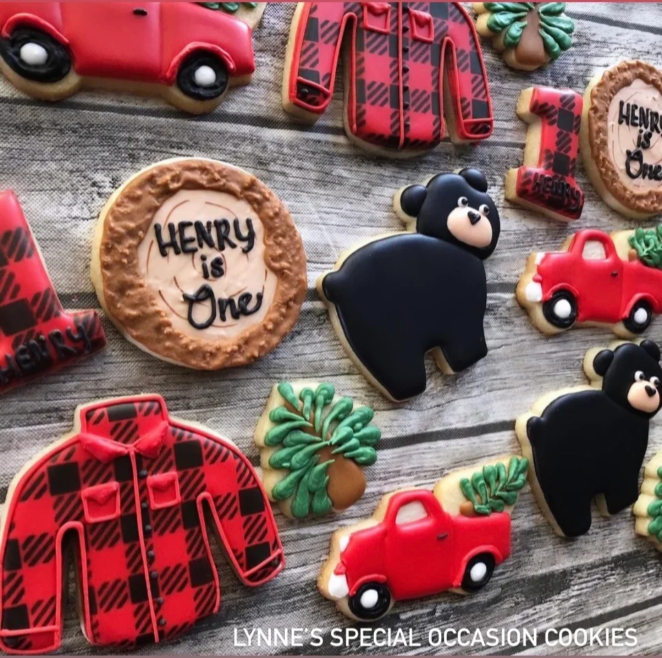 Handmade decorated sugar cookies with a lumberjack birthday theme, featuring red plaid shirts, trucks, bears, pine trees, and a “Henry is One” cookie.