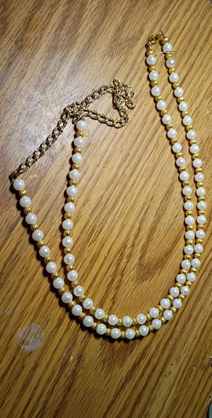 Handmade double‑strand necklace featuring alternating white pearl beads and gold metallic beads with a gold clasp, displayed on a wooden surface.
