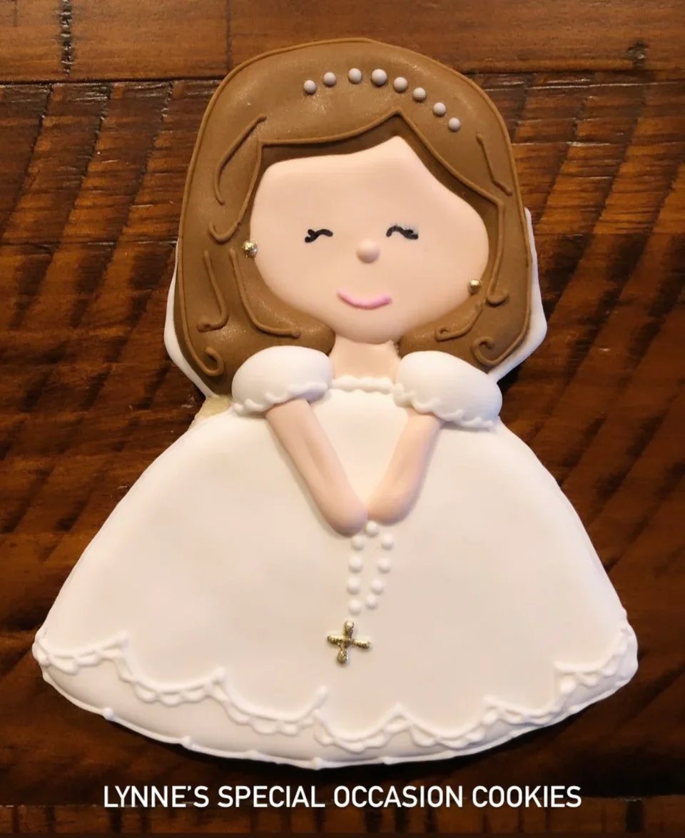 Handmade decorated sugar cookie shaped like a girl in a white First Communion dress holding a rosary, with detailed icing and soft pastel accents.