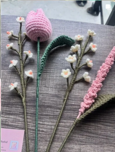 Handmade crochet flower stems including a pink tulip, white blossoms with orange centers, and a textured pink flower, crafted by Meraki The Soul Creativity and displayed on a wooden surface.