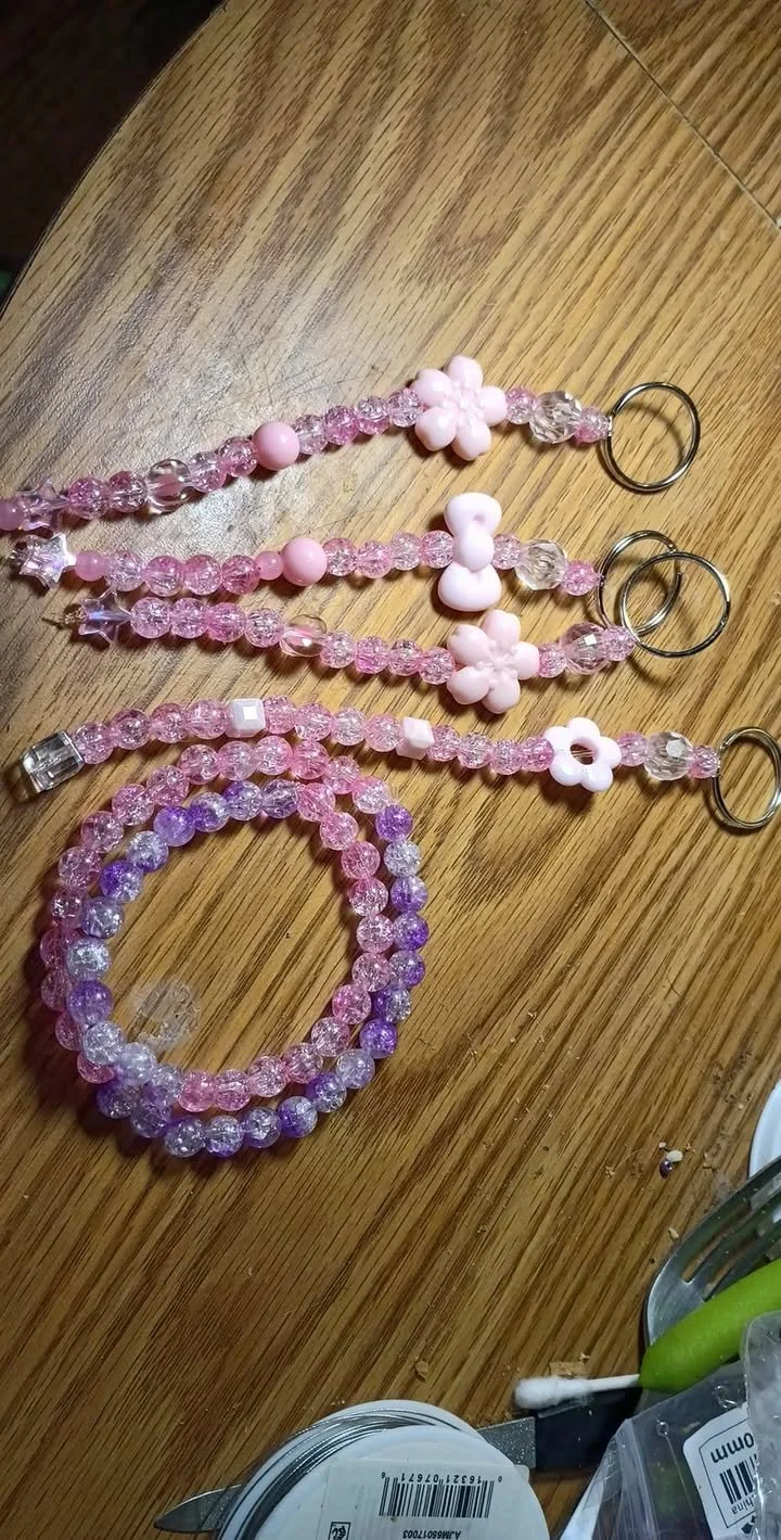 Handmade beaded accessories including pink and clear beaded keychains and a coiled crackle‑bead bracelet, displayed on a wooden surface with jewelry‑making tools.
