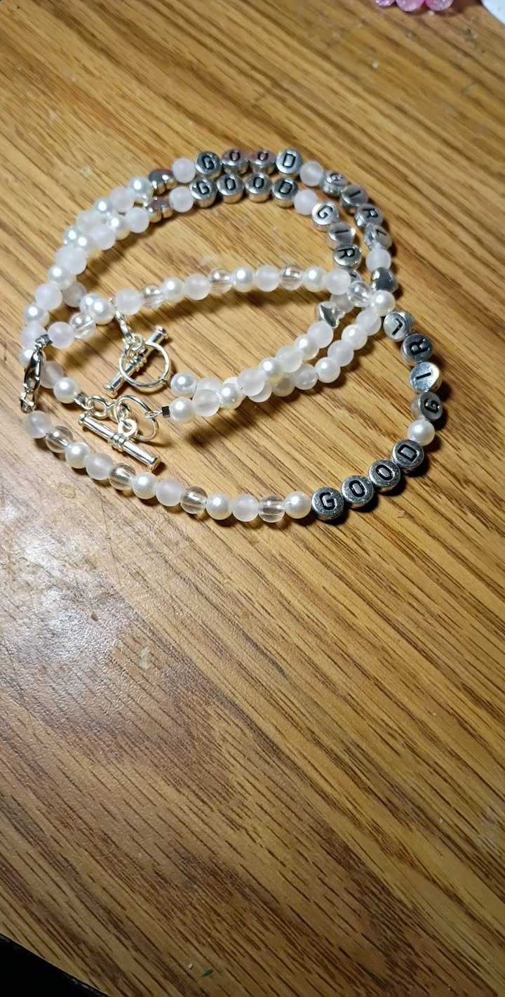 Handmade beaded bracelet with silver and translucent white beads featuring letter beads spelling “GOOD GIRL,” finished with a silver toggle clasp on a wooden surface.