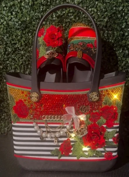 Coordinated embellished handbag and slip‑on shoes featuring red rose embroidery, sequins, gold accents, pearl details, and matching decorative motifs displayed against a leafy background.