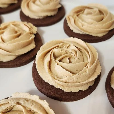 Round chocolate cookies topped with light brown frosting piped in rose‑shaped swirls, arranged on a white surface.