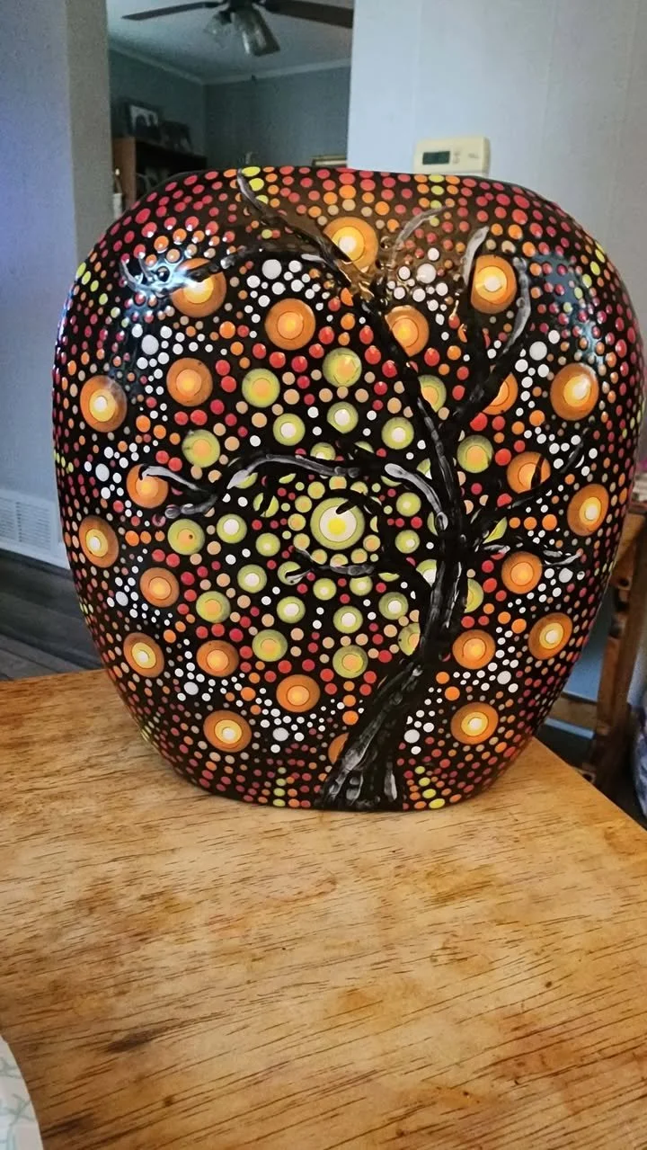 Hand‑painted stone featuring a black tree silhouette surrounded by vibrant mandala dot patterns in red, orange, yellow, and white.