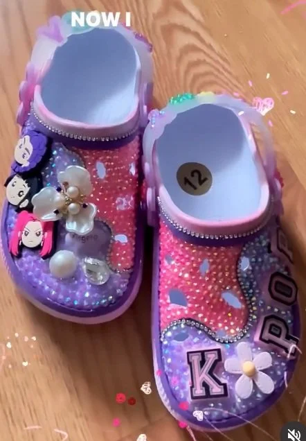 Custom embellished clogs decorated with rhinestones, colorful charms, character accents, and K‑pop themed lettering displayed on a wooden floor.