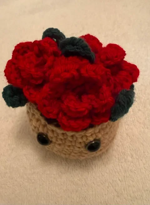 Crocheted plush shaped like a small potted plant with red flowers, green leaves, and a beige pot with button eyes.