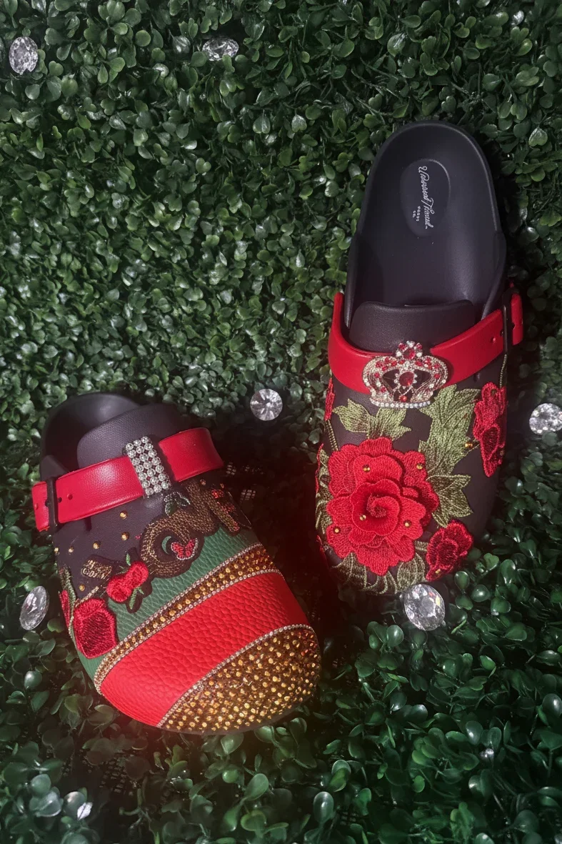 Hand‑embellished slip‑on shoes decorated with embroidered roses, rhinestones, jeweled accents, and gold detailing displayed on a leafy background.