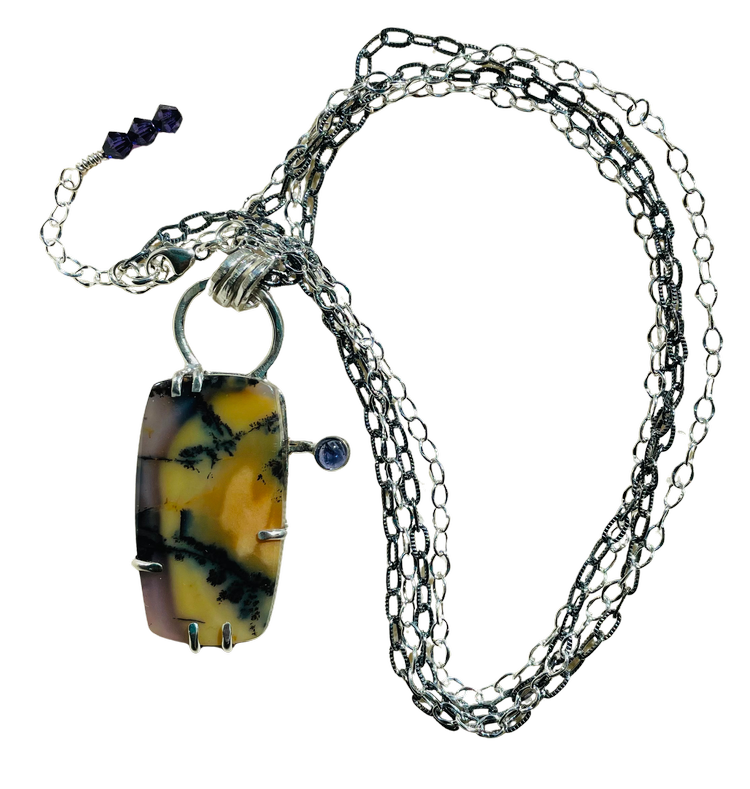 A handcrafted silver necklace featuring a rectangular dendritic gemstone set in a pronged silver frame, accented with a small round blue stone on the side. The pendant hangs from an oval‑link silver chain with dark bead details near the clasp.