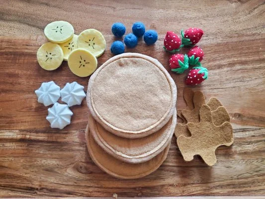 Handmade felt play‑food set featuring pancakes, banana slices, blueberries, strawberries, whipped cream pieces, and a cookie, arranged neatly for imaginative play.