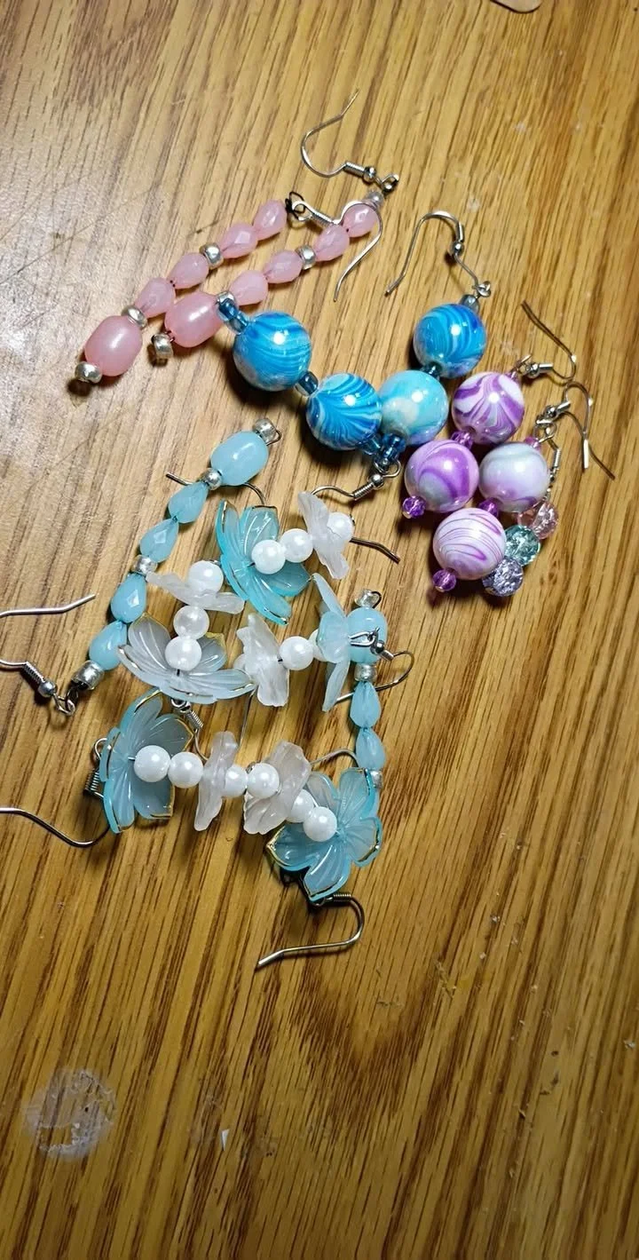Handmade beaded earrings in assorted styles, including pink bead earrings with silver accents, blue and white swirled bead earrings, and light blue floral bead earrings, displayed on a wooden surface.