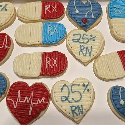 Decorated cookies in heart and capsule shapes with medical‑themed designs, including RX labels, stethoscopes, EKG lines, and “25% RN” lettering arranged in a gift box.