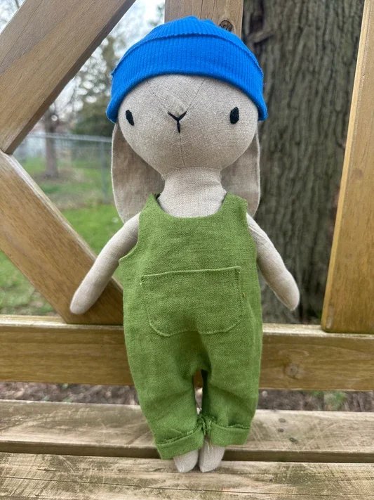Handmade stuffed rabbit toy with stitched facial features, green linen overalls, and a bright blue knit hat, posed outdoors against a wooden backdrop.
