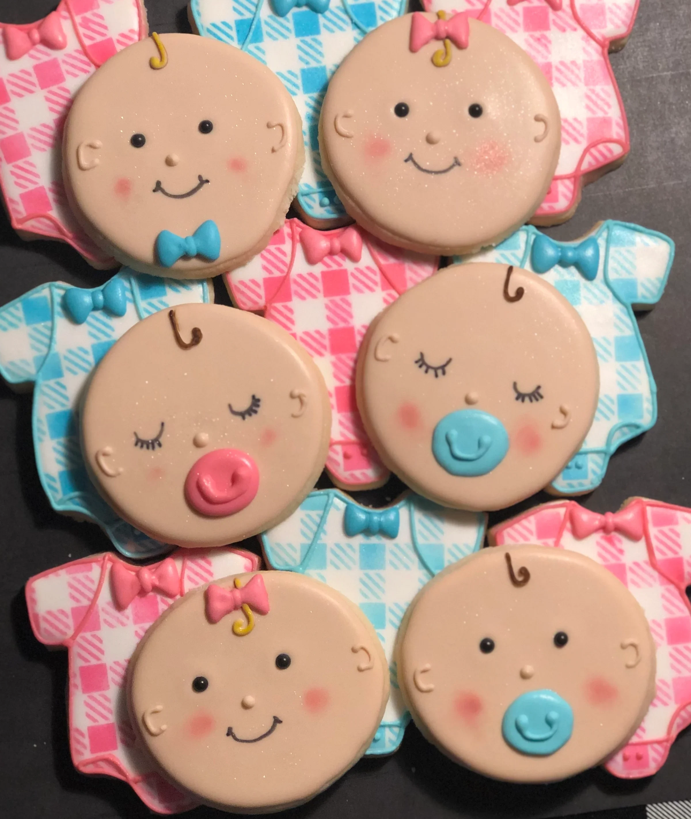 Handmade decorated sugar cookies shaped like baby faces and onesies, iced in pink and blue for baby shower or gender‑reveal celebrations.