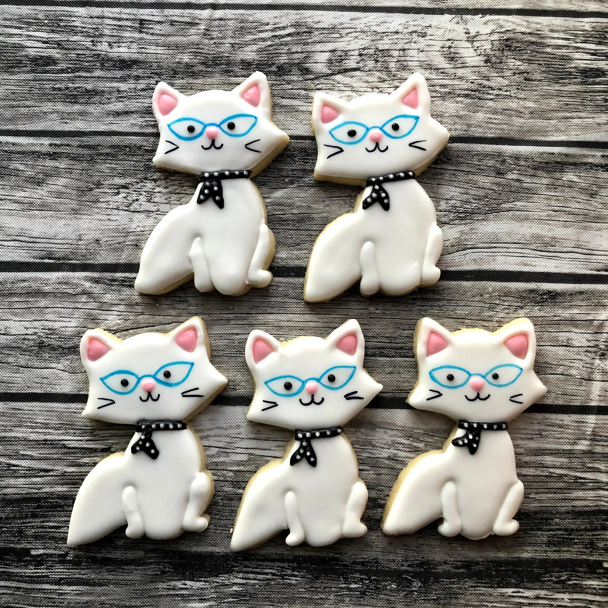 Handmade decorated sugar cookies shaped like white cats wearing blue glasses and black polka‑dot scarves, arranged on a wooden surface.