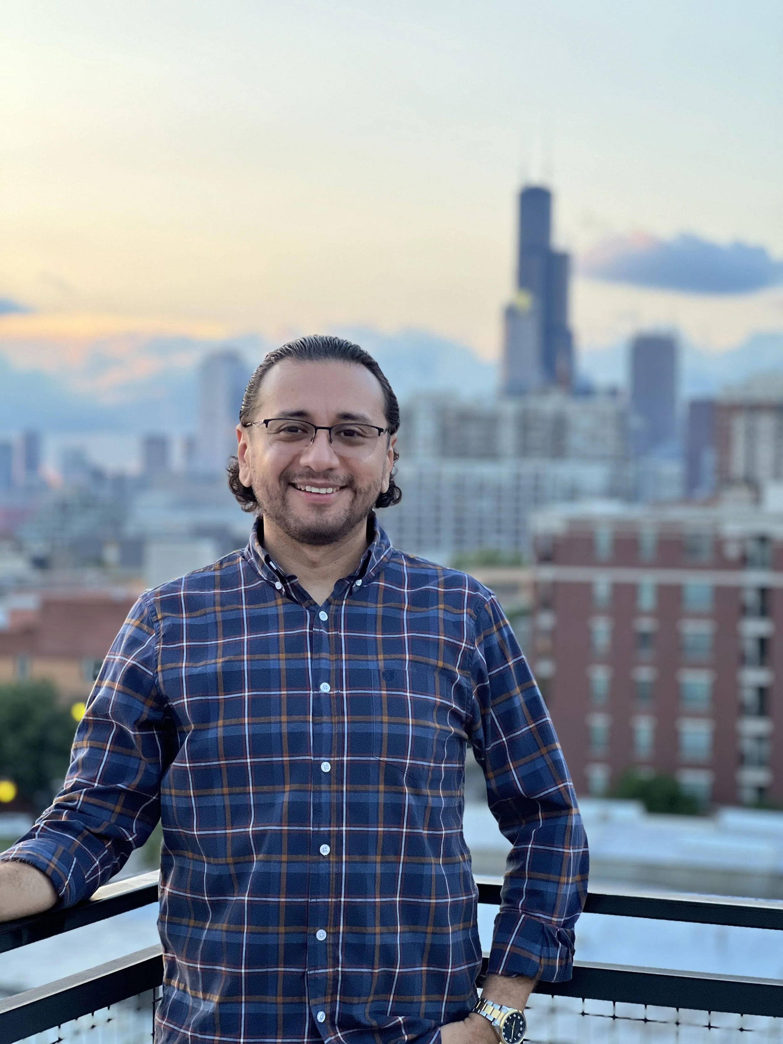 Hombre con gafas y camisa a cuadros en una terraza con vista a la ciudad al atardecer.