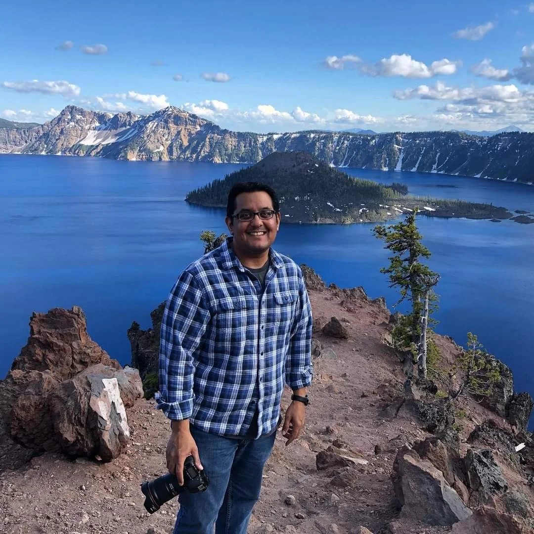 A man standing on rocky ground with a camera in hand, smiling, near a large blue lake surrounded by mountains and trees, under a partly cloudy sky.