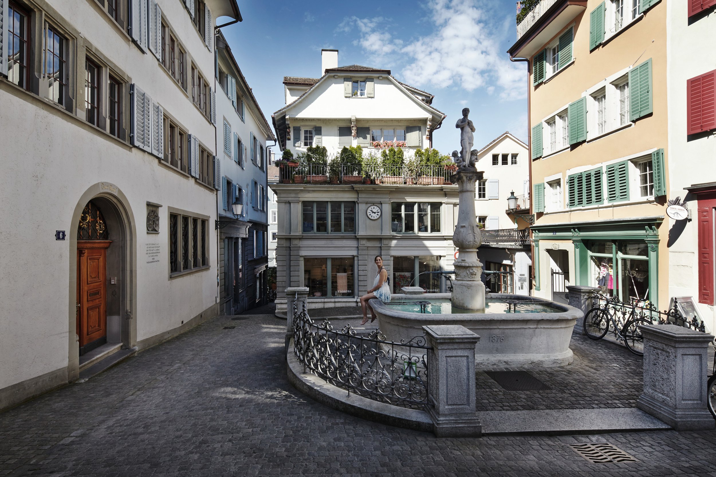 A woman sitting on a fountain in a European-style city square with colorful buildings and a blue sky.