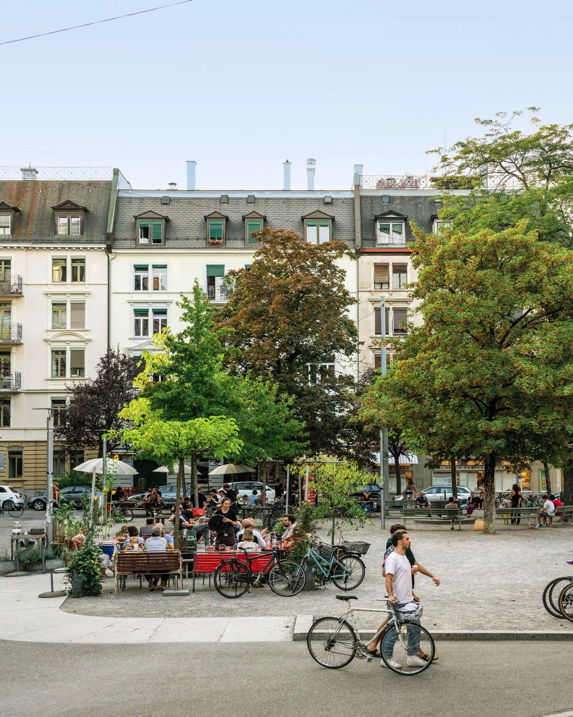 A lively city square with people sitting at outdoor tables, walking, and riding bicycles. Green trees line the area, and multi-story residential buildings with dormer windows can be seen in the background. The scene depicts a warm, social atmosphere.